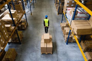 Fulfillment-article–qmsfr92k1y330w3dgcjt4p4ua24mao2hhk5ke99t9a Warehouse worker in warehouse with yellow vest and blue hardhat pulling a cart with boxes
